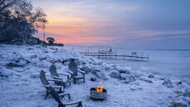 A snow-covered beach with Adirondack chairs, a fire pit, and a dock extending into the water. - Lakefront Cabin Hot Tub, Dock, and Adventure Shed (Sturgeon Bay)