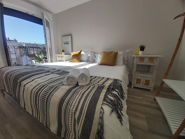 A bedroom with a large bed, a window, a bedside table with a plant, and a small white cabinet.