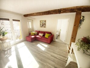 A spacious living room with a red sectional sofa, wooden beams, a balcony with plants, and a view of buildings.