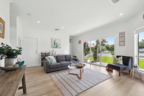 A spacious living room with a grey sofa, a coffee table, and a large window offering a view of the outdoors. - Encounter Retreat (Encounter Bay)