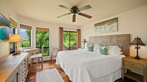 Ein Schlafzimmer mit einem großen Bett, einem Schreibtisch, einem Stuhl, einem Deckenventilator und Blick ins Grüne.