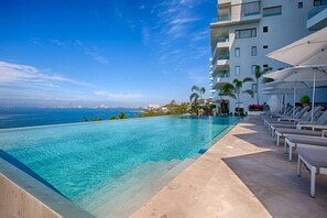 A swimming pool with a view of the ocean, lounge chairs, and a building with balconies. - Oceanfront 3BR | Pools, Spa, Gym & Restaurants (Puerto Vallarta)