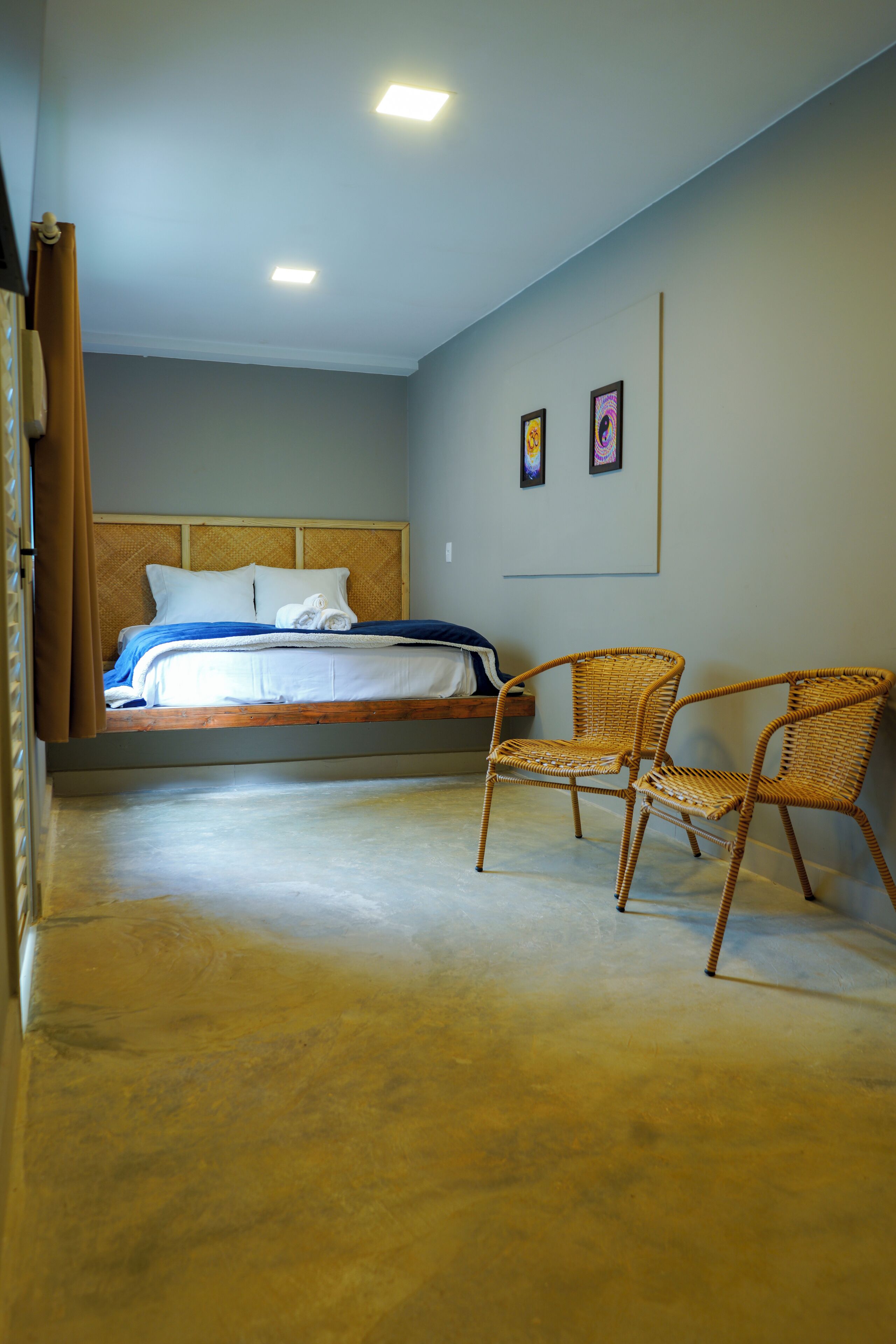 Un dormitorio con una cama, dos sillas de mimbre, un cabecero de madera y dos cuadros en la pared.