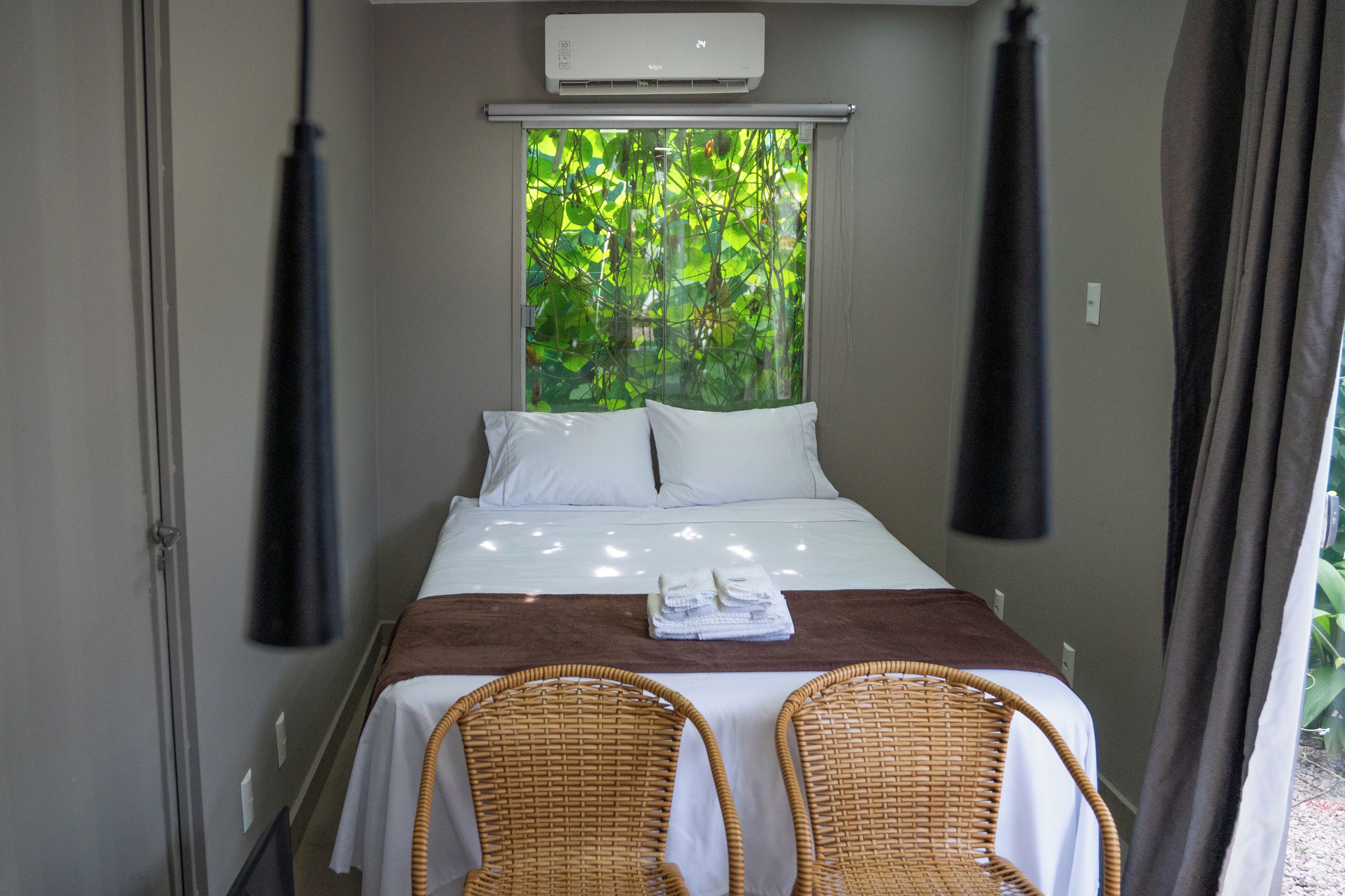 Una camera d'hotel con un letto, due poltrone in vimini, una finestra vista verde e un condizionatore.