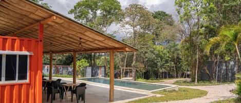 A covered outdoor area with a pool, surrounded by greenery and a fenced garden.