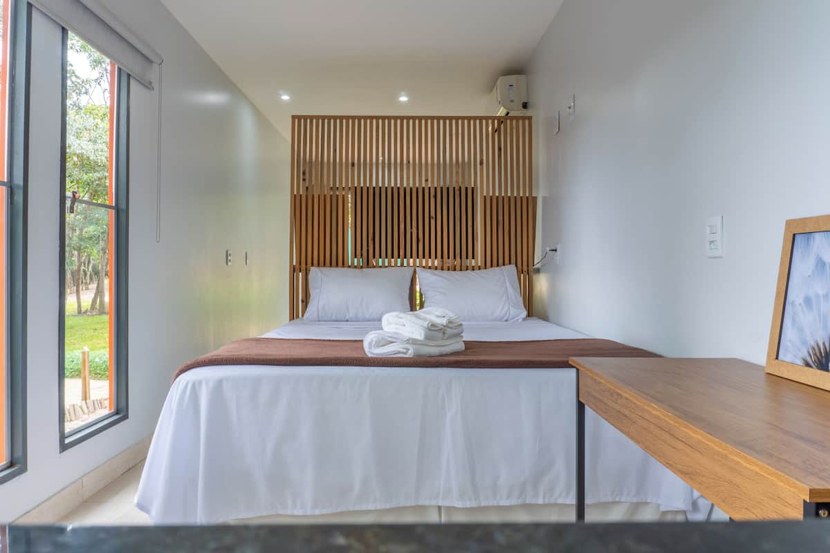 Ein modernes Hotelzimmer mit einem großen Bett, einem Holzkopfschild, einem Nachttisch aus Holz und Blick auf Grünflächen.