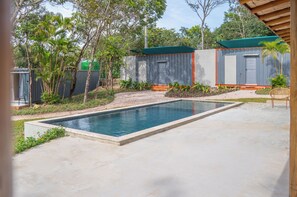 Ein Schwimmbecken mit Betonumrandung, umgeben von einem eingezäunten Bereich mit einem kleinen Gebäude und Palmen.