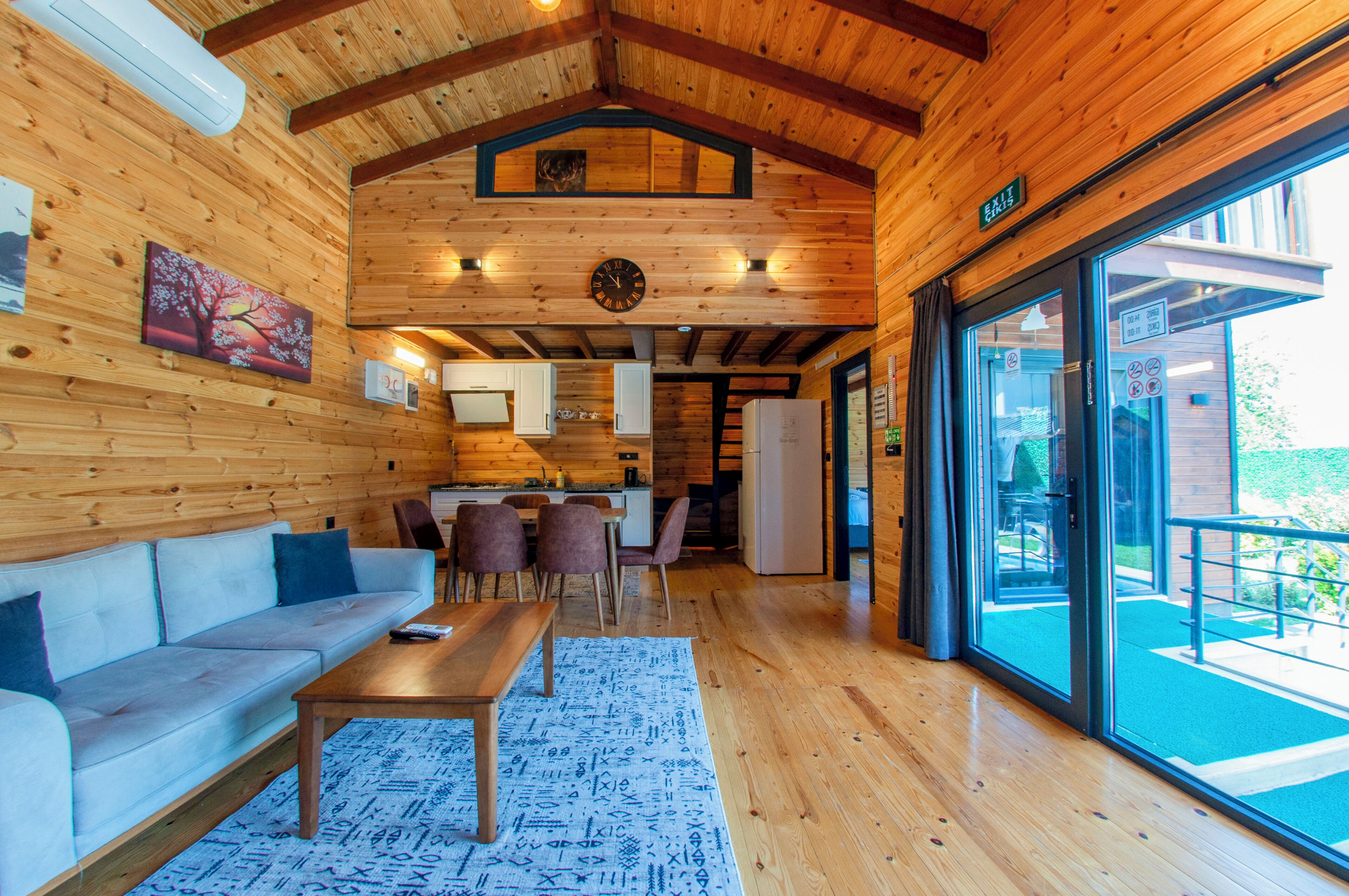 A wooden interior with a sofa, dining table, and chairs. There is a kitchen area with cabinets and a refrigerator. A large window provides a view of the outdoors.