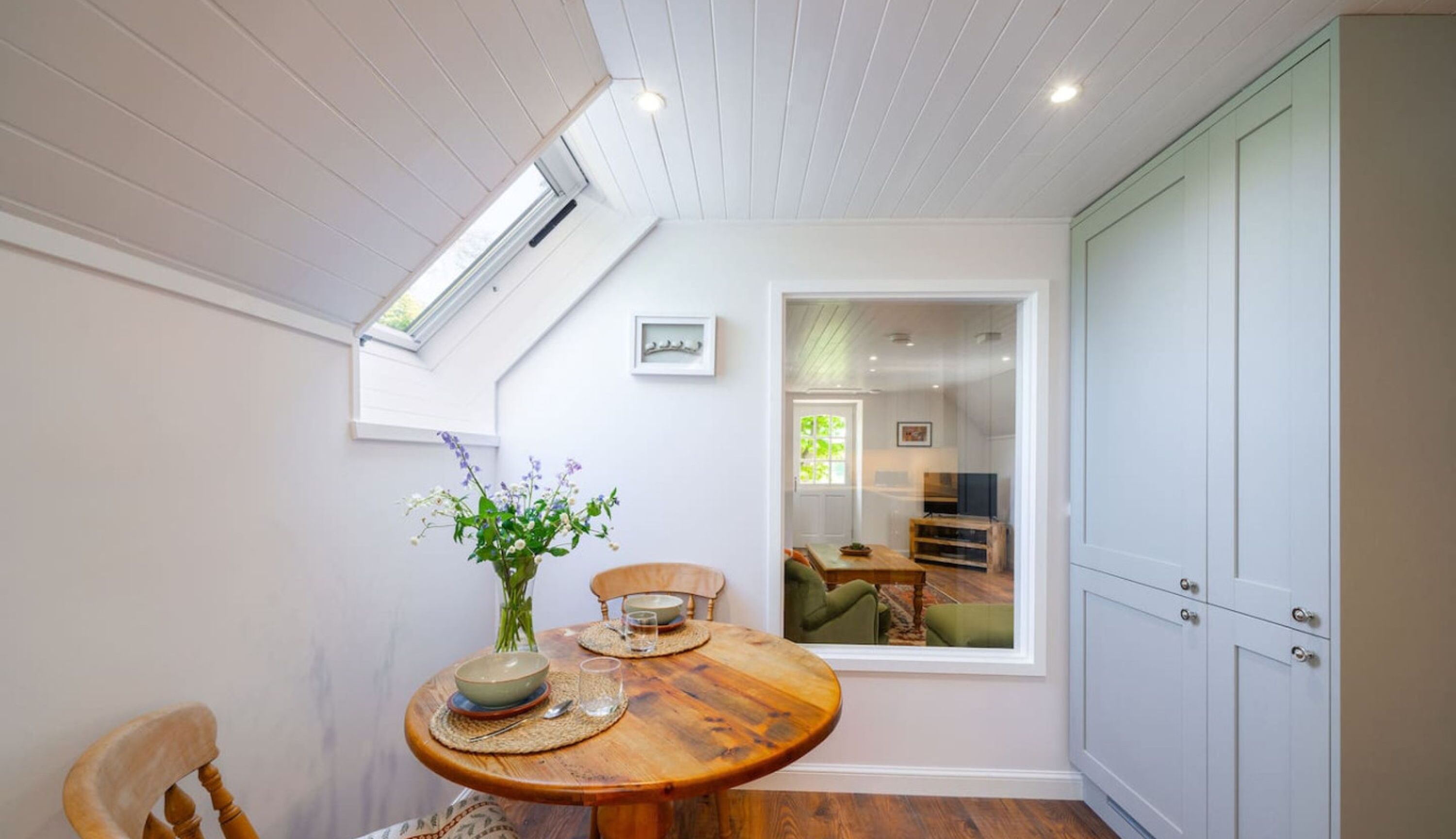 A dining area with a wooden table, chairs, and a vase of flowers. There is a window with a view of another room.