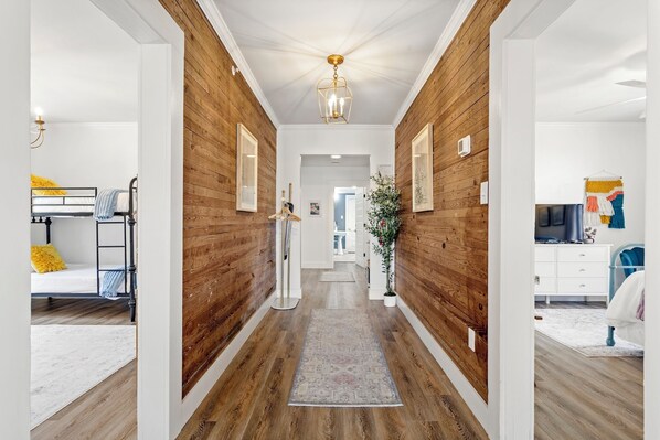 Un pasillo con paredes de madera, una lámpara central y un tapete que conduce a un dormitorio con cama y cómoda.