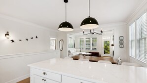 A modern kitchen with white cabinetry, a large island, pendant lighting, and a view to a living area with a white sofa and dining table. - Seabrook's Tori Pines: Hot Tub, Fire Pit, Outdoor Dining, Pet Free! (Pacific Beach)