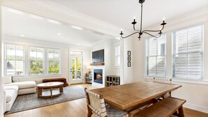 A spacious living room with a wooden dining table, a fireplace, and large windows. - Seabrook's Tori Pines: Hot Tub, Fire Pit, Outdoor Dining, Pet Free! (Pacific Beach)