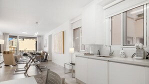Una cocina moderna con armarios blancos, un comedor con mesa de vidrio y vistas a la ciudad a través de amplios ventanales.