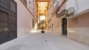 Un estrecho callejón empedrado con un árbol en maceta en el centro, flanqueado por edificios con balcones y unidades de aire acondicionado.