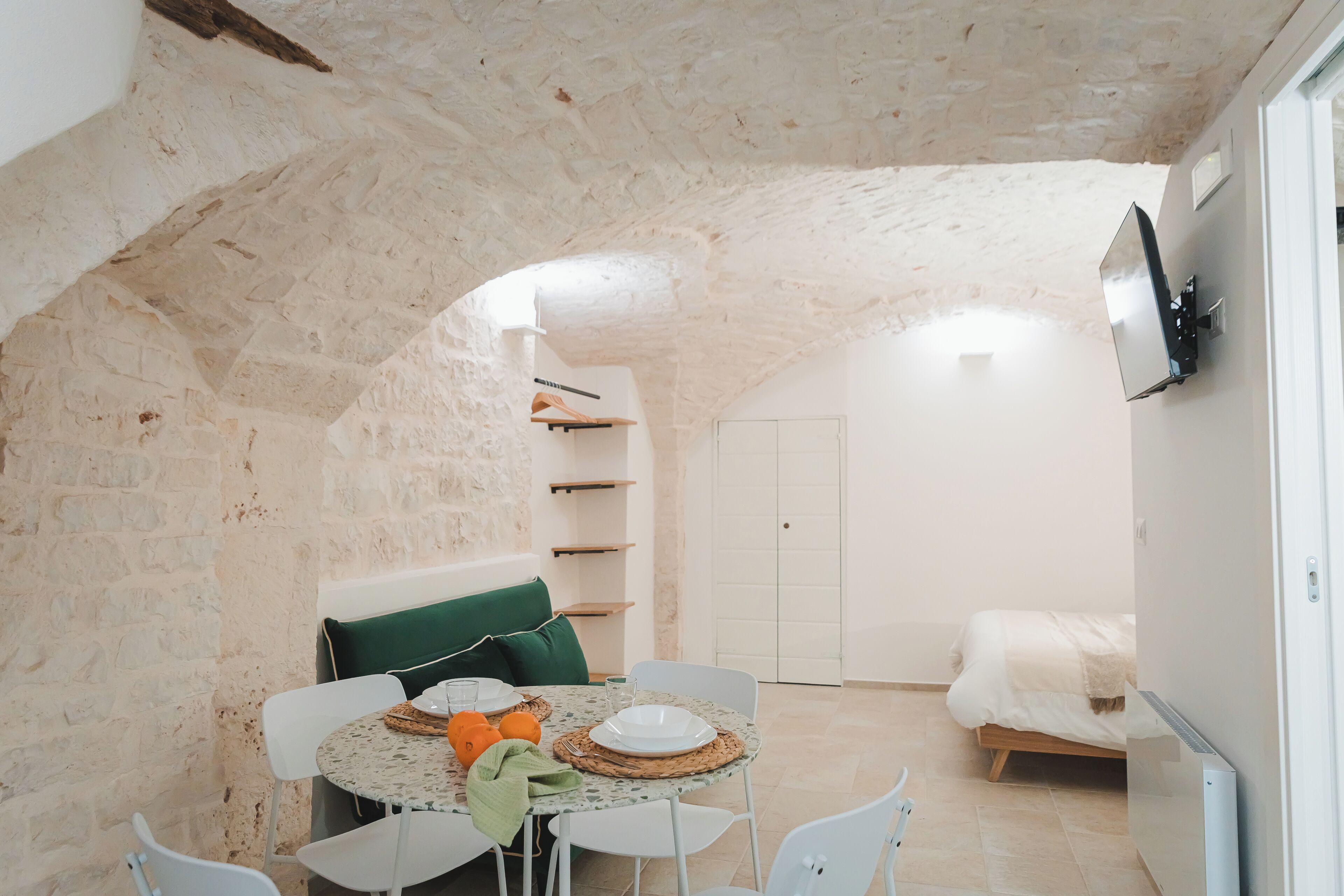Un coin repas accueillant avec une table ronde, des chaises blanches et un canapé vert. Un téléviseur est fixé au mur, et on aperçoit un lit au fond.
