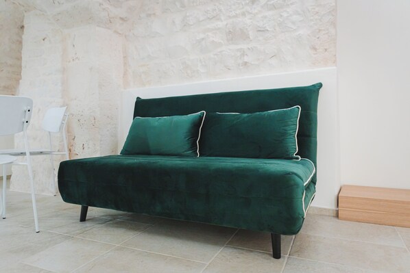 A green velvet sofa with two pillows against a textured wall, a white chair, and a wooden bench in a room with tiled flooring. - Dimore Bianche Santa Chiara  (Putignano)