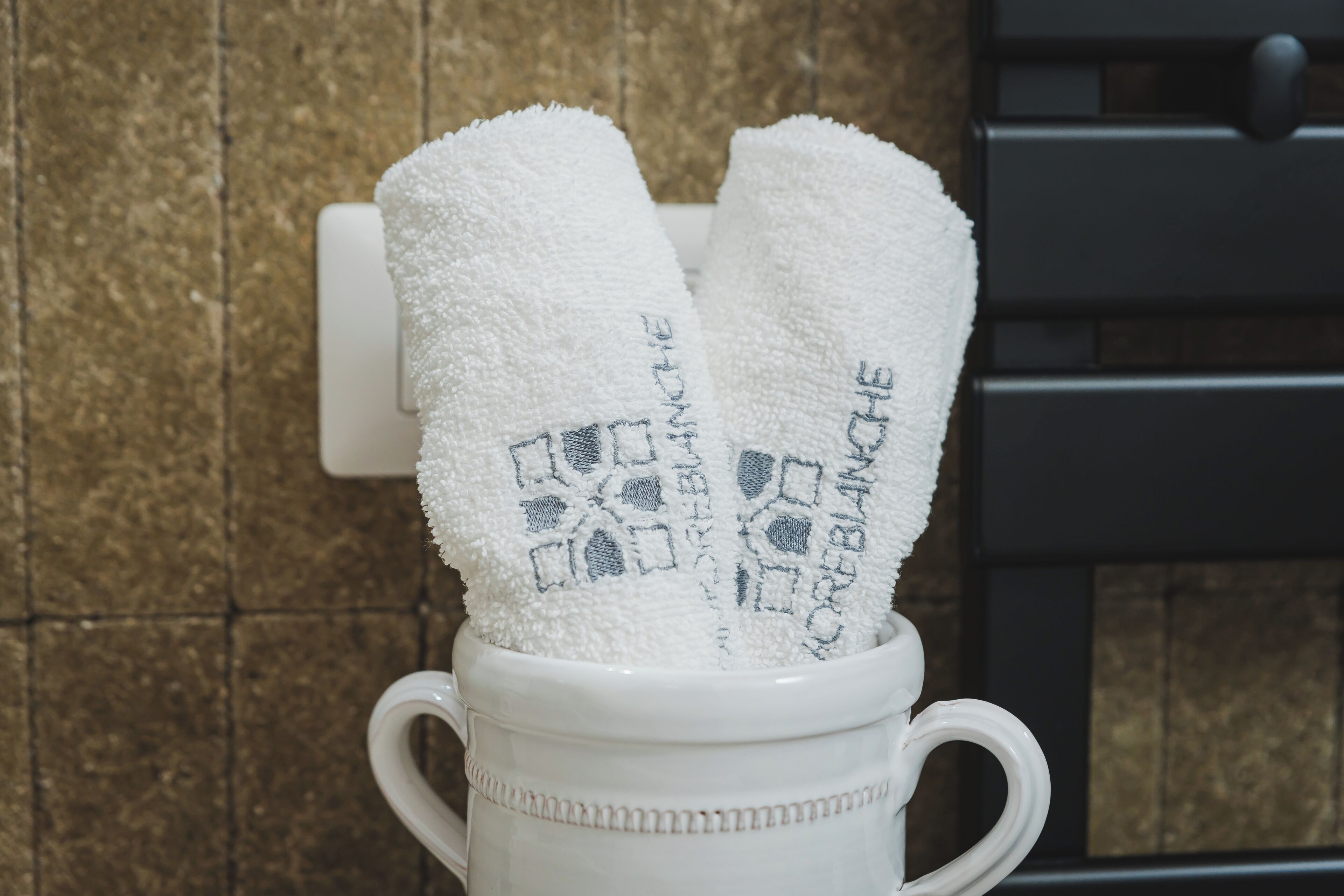 Une tasse en céramique blanche avec des anses en forme de cœur, contenant deux serviettes blanches ornées de motifs brodés.