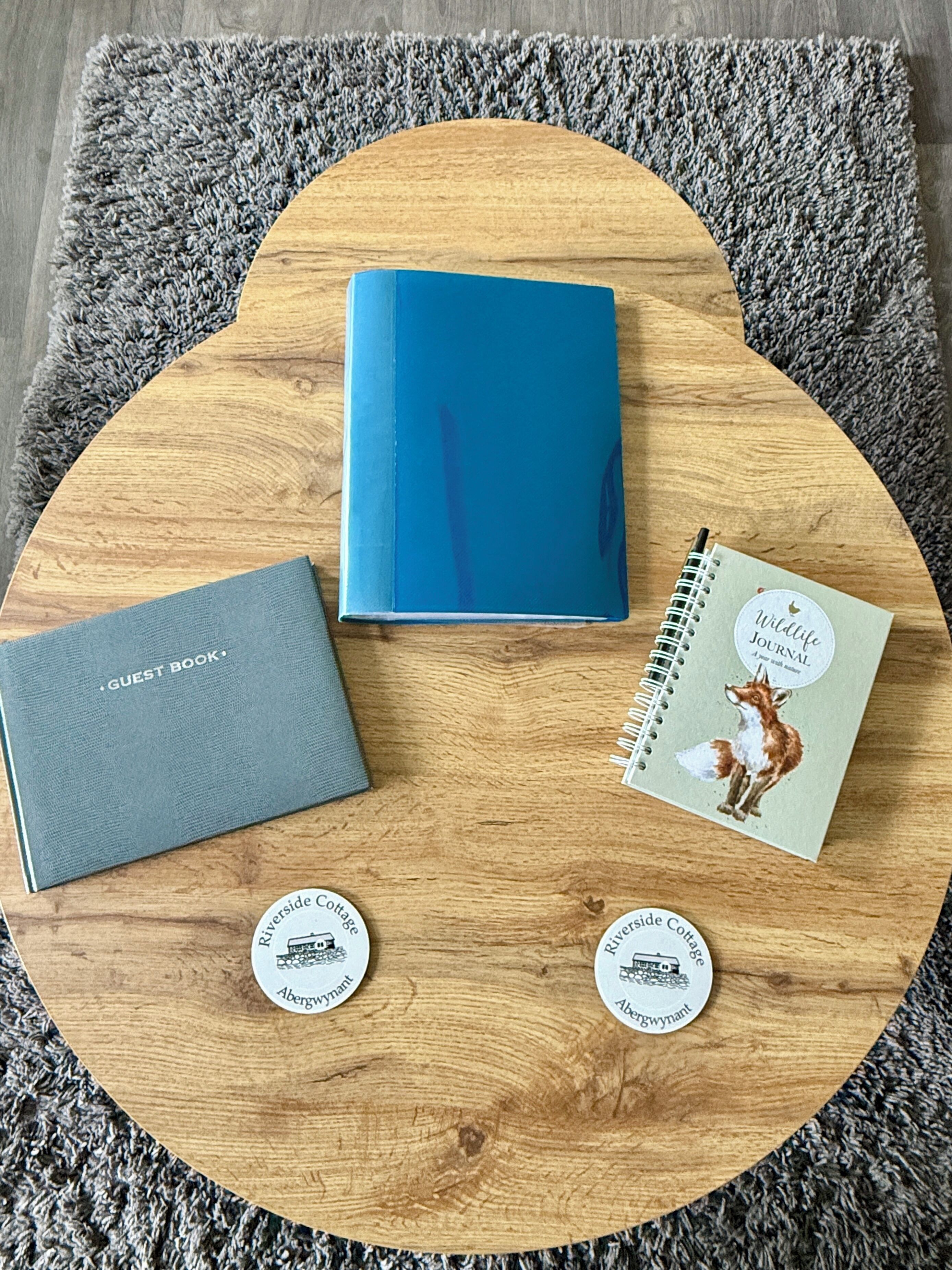 Una mesa de madera con una libreta azul, un libro de visitas gris y dos posavasos circulares con una ilustración de una cabaña.
