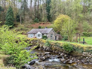 A stone cottage with a thatched roof, surrounded by a lush green landscape and a flowing stream.