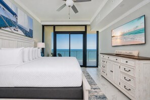 A bedroom with a large bed, a ceiling fan, a view of the ocean through a sliding door, and a painting of a beach scene.