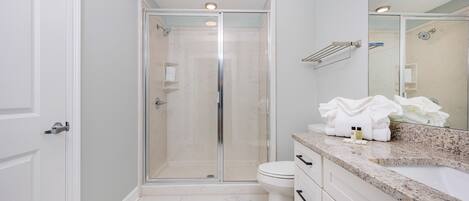 Un baño moderno con cabina de ducha de vidrio, inodoro blanco y encimera de granito con artículos de tocador.