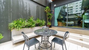 Un espacio exterior moderno con zona de estar que incluye una mesa redonda y sillas, rodeado de plantas en macetas y un muro de cristal con vistas a la ciudad.