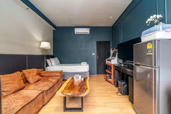 Ett kompakt hotellrum med en soffa, en säng, ett träbord, en liten köksdel med kylskåp och en luftkonditioneringsenhet på väggen.