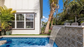 A swimming pool with a waterfall feature, surrounded by tropical plants and a white building with large windows. - 83rd St Villas East 2 Bedroom Villa (Holmes Beach)