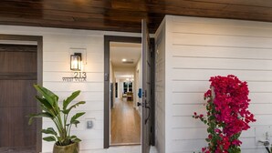 A modern villa entrance with a wooden door, a potted plant, and a vibrant bougainvillea. - 83rd St Villas West 2 Bedroom Villa (Holmes Beach)