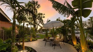 A patio area with a round table and chairs, surrounded by tropical plants and a large umbrella. - 83rd St Villas West 2 Bedroom Villa (Holmes Beach)