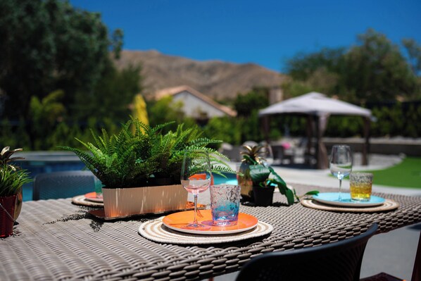 Un área de comedor al aire libre con una mesa de mimbre, plantas en macetas y una vista despejada de una piscina y montañas al fondo.