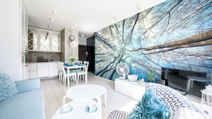 A modern living room with a large tree branch mural, a dining area with white furniture, and a kitchen with white cabinets. - Apartamenty Sun & Snow Nad Zalewem (Dziwnówek)