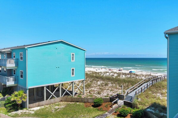 A turquoise building with a balcony, elevated on stilts, overlooking a beach with a wooden walkway.