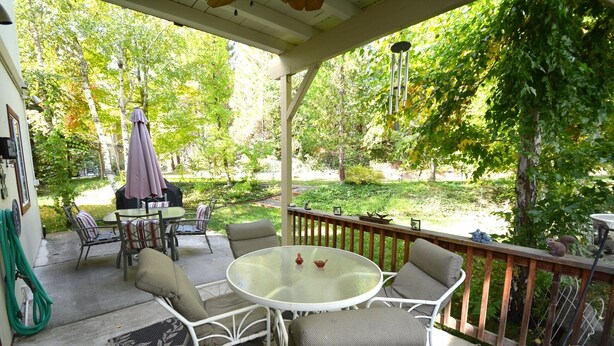 A covered patio with a table and chairs, surrounded by greenery and a wooden railing.