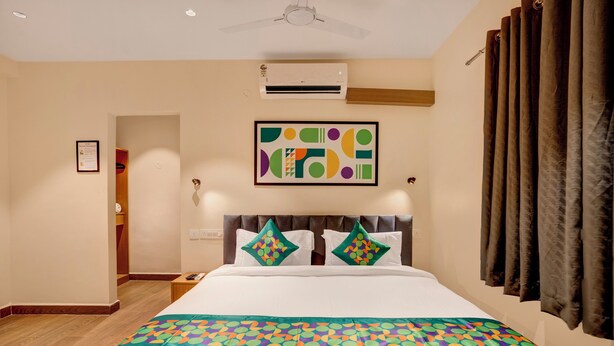 Una habitación de hotel con cama, unidad de aire acondicionado, ventilador de techo, una obra de arte enmarcada y una ventana con cortinas.