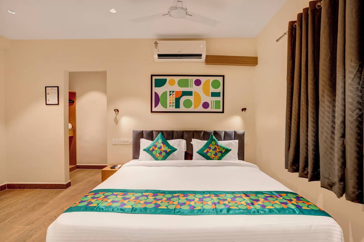 A hotel room with a bed, an air conditioning unit, a ceiling fan, a framed artwork, and a window with curtains.