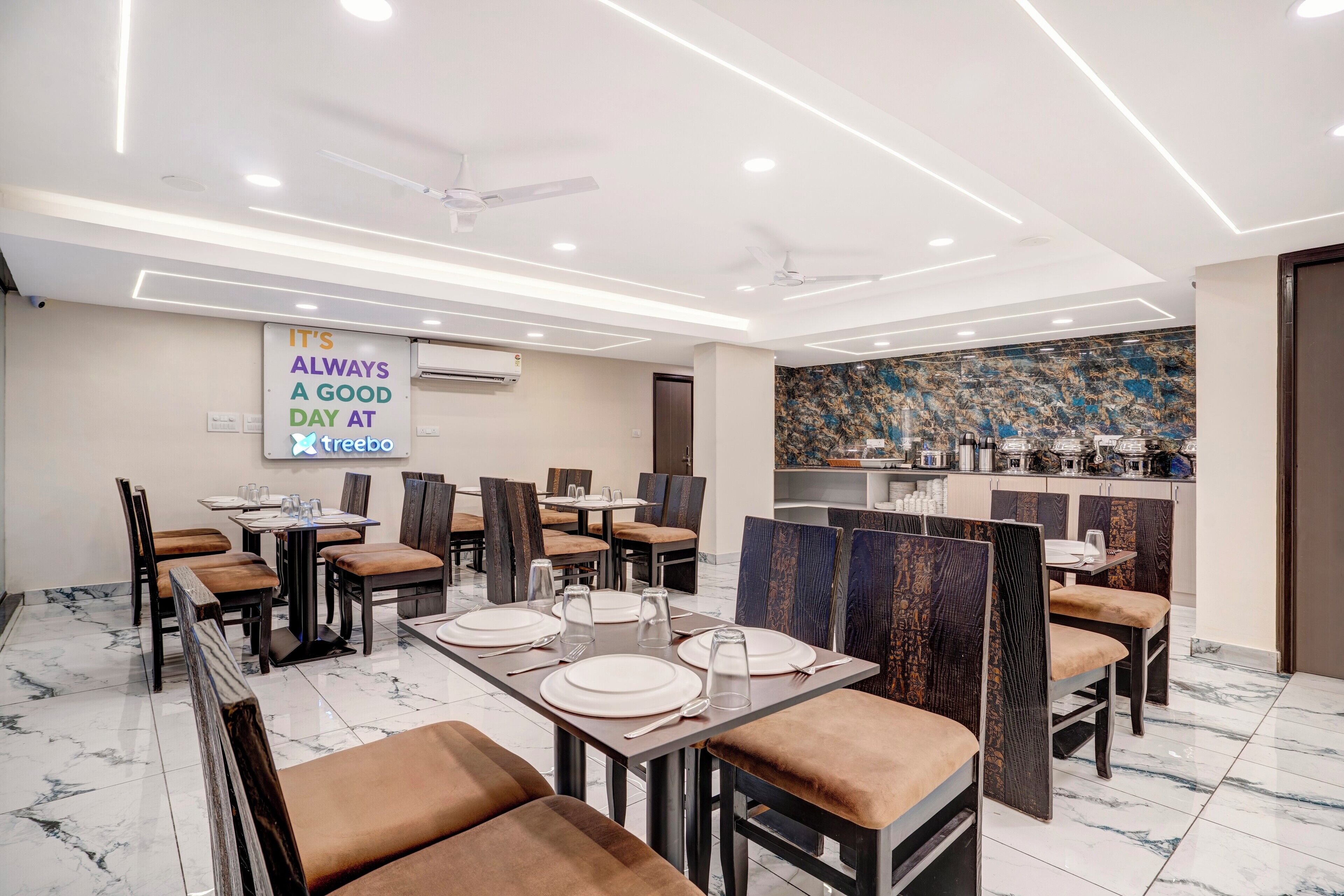 A modern dining area with tables set for four, ceiling fans, and a kitchen in the background.