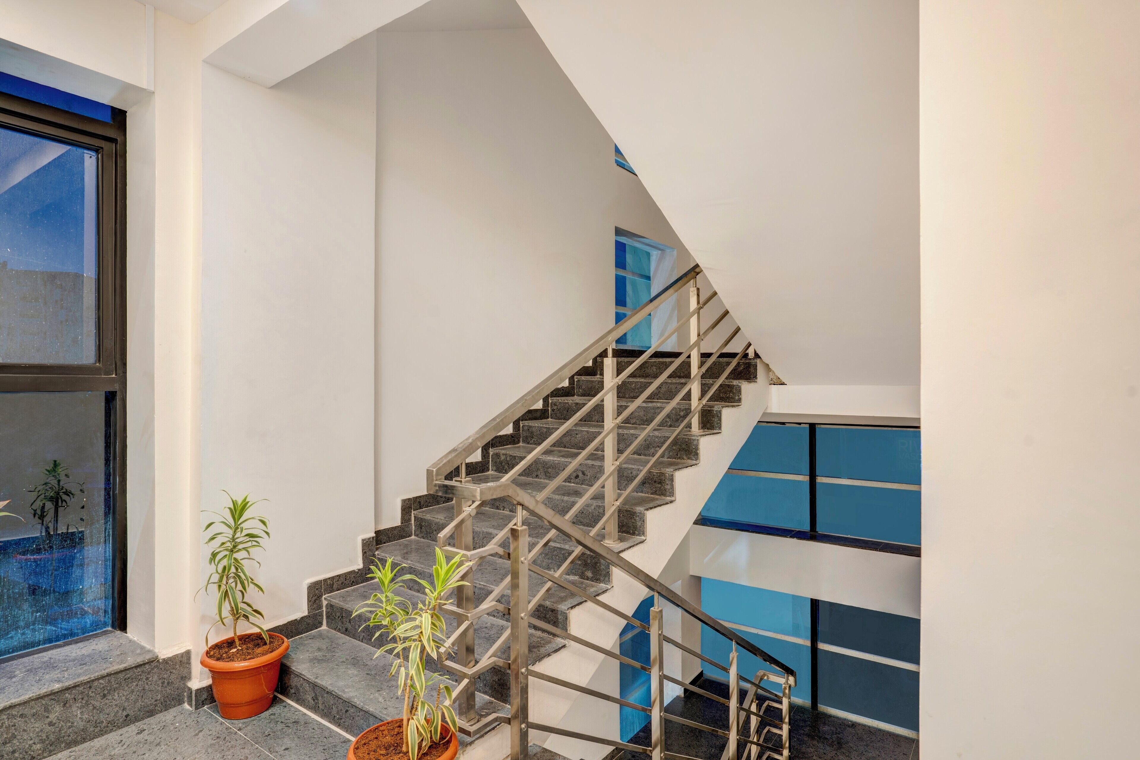 Un escalier moderne avec des rampes en métal, des plantes en pot et une vue sur l’extérieur grâce à une grande fenêtre.
