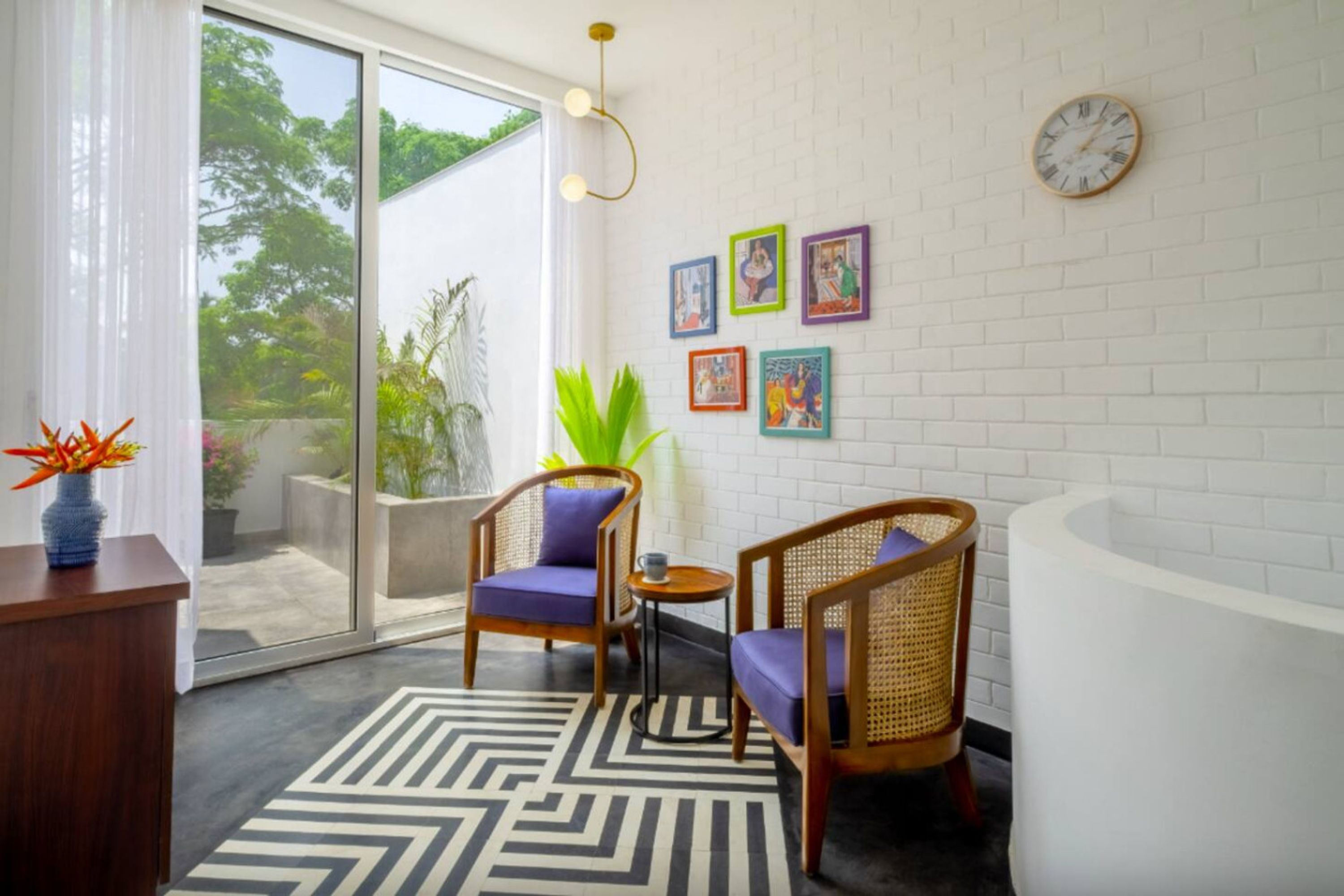 Un bagno moderno con una vasca da bagno bianca, due poltrone in vimini con cuscini viola, un tappeto a motivi bianchi e neri e una parete con una sveglia e quadri colorati.