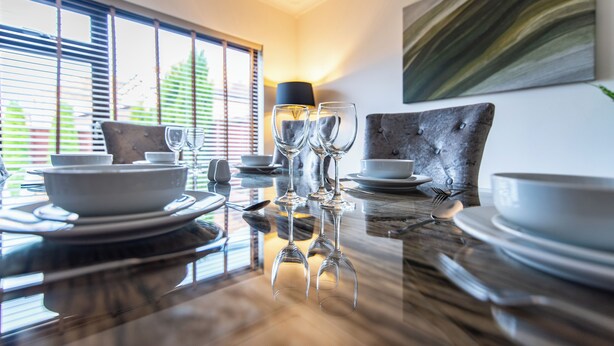 Ένα τραπέζι φαγητού στρωμένο με πιάτα, μπολ και ποτήρια κρασιού, με ένα φωτιστικό και έναν πίνακα στον τοίχο στο βάθος.