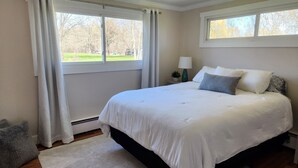 A bedroom with a large bed, white curtains, a small bedside table with a lamp, and a view of trees through the window. - Charming 3-bedroom ranch with a covered back patio, in peaceful Grand Island (Grand Island)