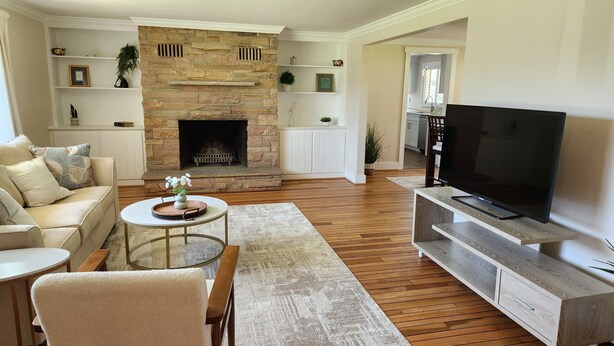 A living room with a stone fireplace, a sofa, a coffee table, a TV, and a wooden floor. - Charming 3-bedroom ranch with a covered back patio, in peaceful Grand Island (Grand Island)