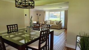 A dining area with a glass-top table and wooden chairs, a sofa with cushions, and a view of a building through a window. - Charming 3-bedroom ranch with a covered back patio, in peaceful Grand Island (Grand Island)