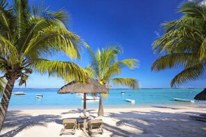 Ein Strand mit Palmen, einem Strohdachschirm und Liegestühlen.