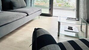 A modern living room with a grey sofa, a black chair, and a glass coffee table. A large sliding glass door leads to a balcony with a mountain view. - Comfy Vista Apartment (Kalamata)