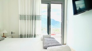 A modern hotel room with a large bed, a flat-screen TV mounted on the wall, and a view of mountains through the window. - Comfy Vista Apartment (Kalamata)