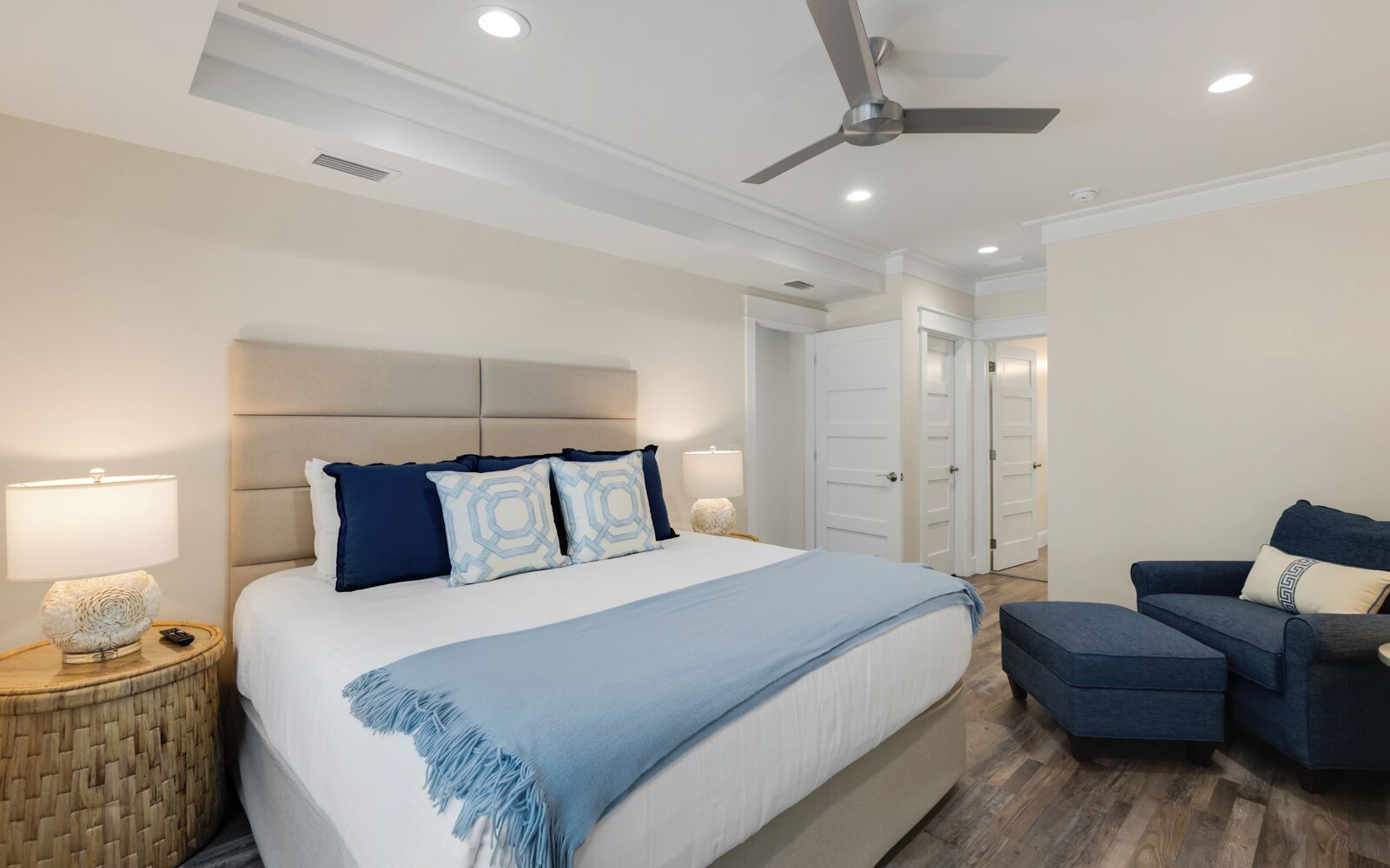 A modern bedroom with a large bed, bedside lamps, a ceiling fan, and a seating area with a sofa and ottoman.