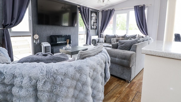 A modern, well-lit room with a grey sofa set, a flat-screen TV mounted on the wall, and a large window with curtains. - 155 Woodland Hall (Ruthin)