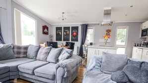 A modern living room with a grey sectional sofa, a smaller grey sofa, and a coffee table. There is a kitchen area with white cabinets and a dining table. The room has large windows and a ceiling fan. - 155 Woodland Hall (Ruthin)