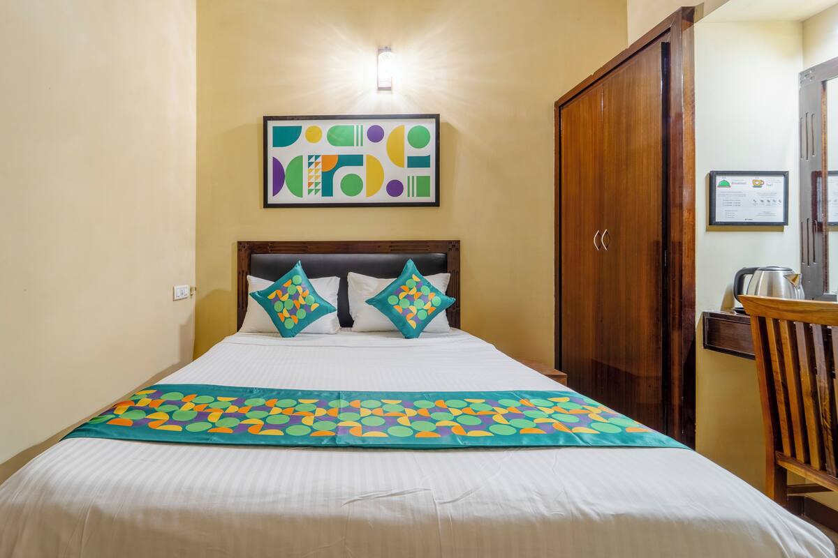 Un lit bien fait, agrémenté d’une couvre-lit aux couleurs vives, une tête de lit ornée de coussins décoratifs, une armoire en bois et une œuvre d’art abstraque encadrée accrochée au mur.